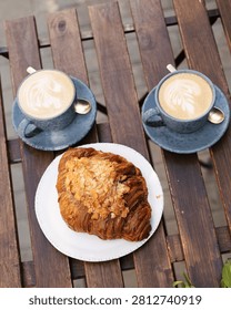 Two ceramic cups filled with latte with art, and a plate with an almond-topped croissant on a slatted wooden outdoor table.