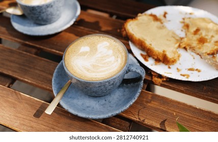 Two ceramic cups filled with latte with art, and a plate with a partially eaten croissant on a slatted wooden outdoor table.
