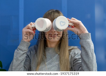 Two ceramic teacups with hearts painted on the bottom in the hands of a smiling woman. A pretty lady is fooling around by covering her eyes with cups. Selective focus in the drawing.