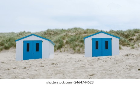 Two charming beach huts with vibrant blue doors stand out amidst the sandy shores of Texel, inviting visitors to relax and enjoy the seaside views. De Koog beach Texel - Powered by Shutterstock