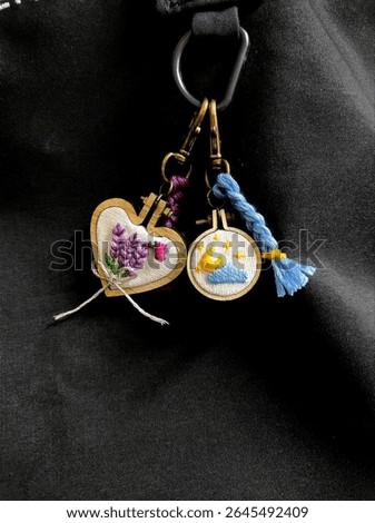 Two charming handmade embroidered keychains, one heart-shaped with lavender, the other round with moon and stars. Features tiny wooden hoops, braided threads, and brass clasps on a dark background. 