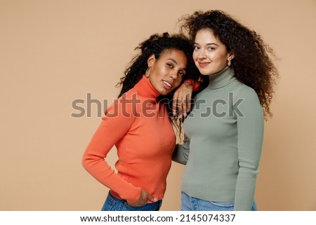 Two charming young curly black women friends 20s wear casual shirts clothes posing hugging looking camera isolated on plain pastel beige background studio portrait. People emotions lifestyle concept