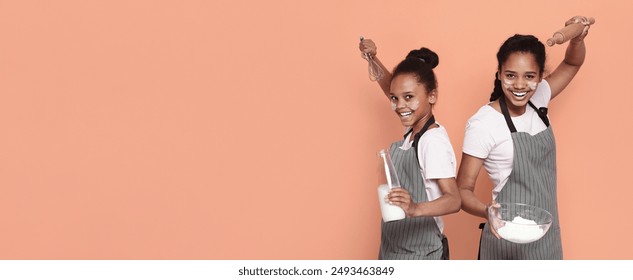 Two cheerful african american sisters with aprons on ready for baking, having flour marks on their cheeks, holding milk, flour and dough spin, pink studio background - Powered by Shutterstock