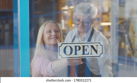 Two Cheerful Aged Small Business Owners Smiling While Putting Open Sign At Cafe Entrance Door. Senior Couple Entrepreneurs Opening New Restaurant. Small Business And Retirement Concept