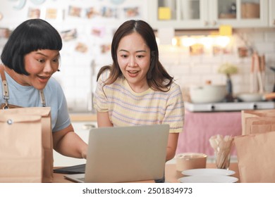 Two cheerful Asian chefs share a joyful moment as they look at a laptop in a cozy kitchen. Surrounded by takeaway bags and delicious food items, they exhibit a sense of accomplishment and happiness - Powered by Shutterstock