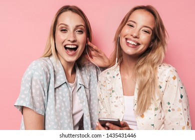 Two cheerful attractive blonde girls wearing summer outfit standing isolated over pink background, listening to music with earphones, holding mobile phone