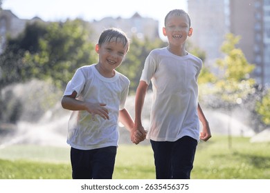 Two cheerful boys are smiling while walking through a park on sunny day, enjoying the summer weather. They are wearing casual wet clothes, suggesting fun activities like water games or sprinkler play.