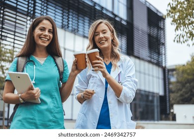 Two cheerful Caucasian female healthcare professionals enjoy a coffee break outdoors, one holding a digital tablet, reflecting a blend of technology use and leisure in their medical roles. - Powered by Shutterstock