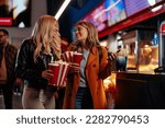 Two cheerful Caucasian girls are in the movie theater having a nigh out, holding tickets, popcorn and beverages in their hands.