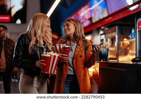 Two cheerful Caucasian girls are in the movie theater having a nigh out, holding tickets, popcorn and beverages in their hands.
