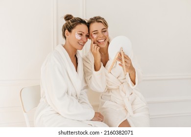 Two cheerful caucasian girls with patches under their eyes grimace looking in mirror sitting indoors. Blondes wear white bathrobes after spa treatments. Skin care and health concept