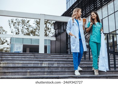 Two cheerful Caucasian women in healthcare attire enjoy a light conversation while descending stairs outside a medical facility, a coffee cup in one's hand signals a relaxed end to their workday. - Powered by Shutterstock