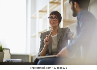 Two cheerful colleagues smiling while discussing something in modern office, focus on stylish businesswoman wearing creative haircut and glasses