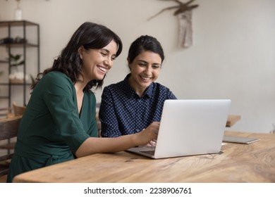 Two cheerful diverse office workers women cooperating on project, sitting at work desk with laptop, looking at screen, smiling, laughing. Female mentor teaching Indian student, pointing at monitor
