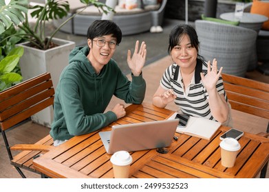 Two cheerful, friendly young Asian male and female friends are sitting at a table outdoors and waving their hand to the camera. friendship, teamwork, companion - Powered by Shutterstock