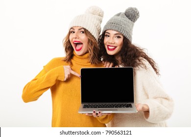 Two Cheerful Girls In Sweaters And Hats Standing Together While Showing Blank Laptop Computer Screen And Looking At The Camera Over White Background