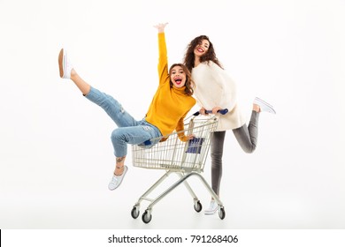 Two cheerful girls in sweaters having fun together with shopping trolley while looking at the camera over white background - Shutterstock ID 791268406