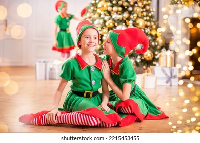 Two Cheerful Happy Girls-friends In Costumes Of Santa's Elves On Background Of Christmas Tree And Other Children