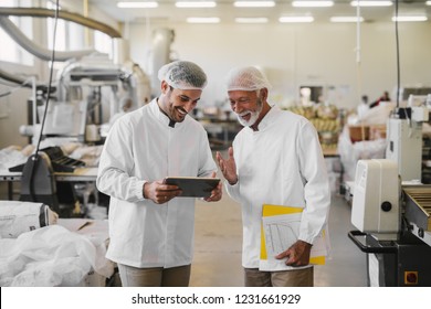 Two Cheerful Male Colleagues In Sterile Clothes Standing In Food Factory And Looking At Sales Documents On Digital Tablet.