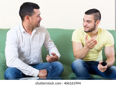 Two Cheerful Men Talking While Sitting On The Couch In The Living Room. Focus On The Right Man