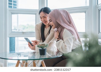 Two cheerful multi ethnic young women looking at phone together while sitting indoors in cafe, talking and laughing - Powered by Shutterstock