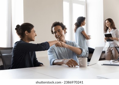 Two cheerful office friends celebrating business task completion, successful project startup, job promotion, sitting at shared workplace, giving fist bumps, talking, smiling, laughing - Powered by Shutterstock