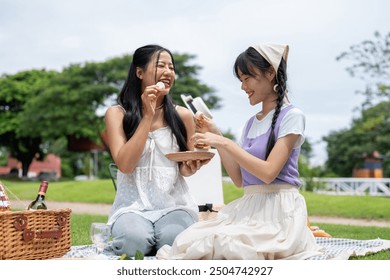 Two cheerful and pretty Asian women are enjoying a picnic together in a green park on a bright day, eating macarons and snacks on a picnic blanket. leisure, hobby, friendship - Powered by Shutterstock