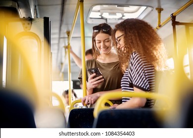 Two cheerful pretty young women are standing in bus   looking at the phone   smiling while waiting for bus to take them to their destination 