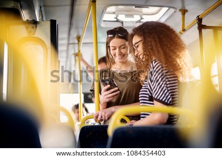 Two cheerful pretty young women are standing in a bus and looking at the phone and smiling while waiting for a bus to take them to their destination.