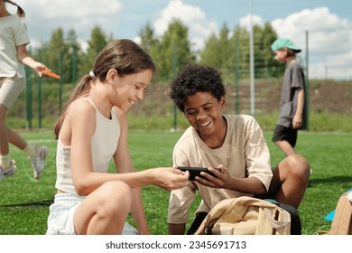 Two cheerful schoolchildren laughing at funny online video in cellphone while sitting on green football field or stadium in front of camera - Powered by Shutterstock