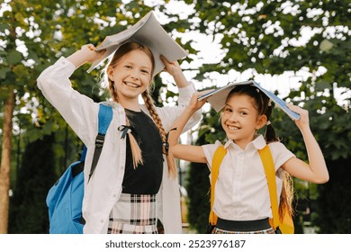 Two cheerful schoolgirls holding books over their heads and smiling outdoors, enjoying their time before going back to school - Powered by Shutterstock