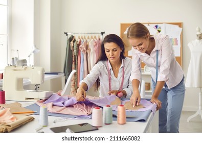 Two cheerful seamstresses in sewing workshop discuss fabric, pattern and sketch during production process. Tailors help each other in friendly way to get great result. Concept of tailoring.