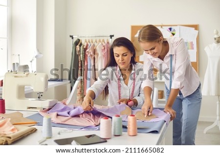 Two cheerful seamstresses in sewing workshop discuss fabric, pattern and sketch during production process. Tailors help each other in friendly way to get great result. Concept of tailoring.