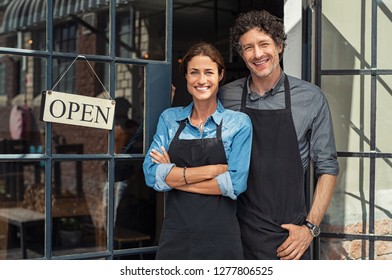 Two cheerful small business owners smiling and looking at camera while standing at entrance door. Happy mature man and mid woman at entrance of newly opened restaurant with open sign board. - Shutterstock ID 1277806525