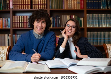 Two Cheerful Students Perform Together The Project Sitting In The Library Against The Backdrop Of Bookcases. Student Science. High Quality Photo