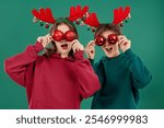 Two cheerful teenagers - a boy and a girl, dressed in red and green clothes and reindeer antlers, are having fun with Christmas tree balls. Merry Christmas and Happy New Year!