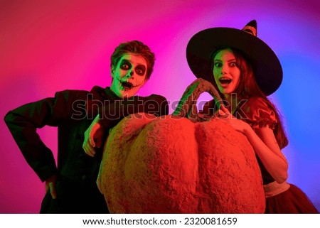 Two cheerful teenagers in costumes of a skeleton and a witch posing together with a big pumpkin. Halloween party. Studio portrait with mixed colorful lighting.