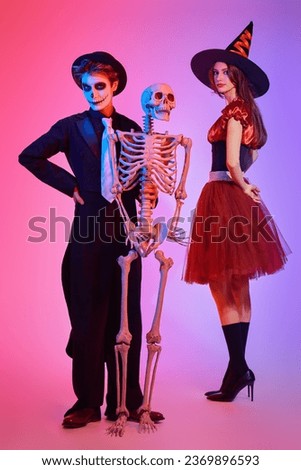 Two cheerful teenagers in costumes of a skeleton and a witch posing together with a skeleton. Full-length studio portrait with mixed colorful lighting. Halloween party.
