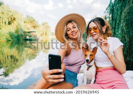 Two cheerful woman friends resting outdoor on lake pier with dog, making photo, selfie