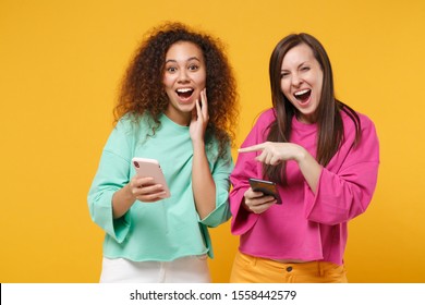 Two Cheerful Women Friends European African American Girls In Pink Green Clothes Posing Isolated On Yellow Background. People Lifestyle Concept. Mock Up Copy Space. Using Mobile Phone, Typing Message