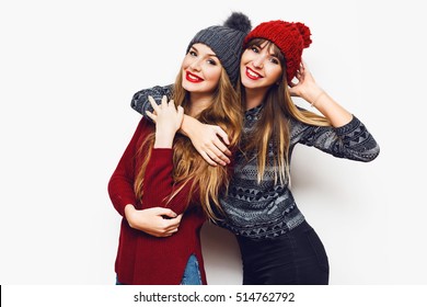 Two  Cheerful Young Friends In Perfect Mood Having Time Together On White Isolate Background. Wearing   Trendy Winter Sweater And Knitted Red And Grey Hat . Positive And Friendship Concept. 