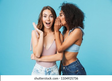 Two cheerful young girls dressed in summer clothes whispering secrets isolated over blue background
