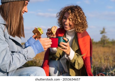 Two Cheerful Young Girls Having Picnic Outdoors During Sunny Autumn Day. Happy Girlfriends Spend Fall Weekend In Countryside Eating Sandwiches And Drinking Hot Tea Or Coffee With Happy Relaxed Smiles