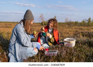 Two Cheerful Young Girls Having Picnic Outdoors During Sunny Autumn Day. Happy Girlfriends Spend Fall Weekend In Countryside Eating Sandwiches And Drinking Hot Tea Or Coffee With Happy Relaxed Smiles