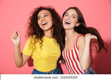 Two Cheerful Young Girls Wearing Summer Clothing Standing Isolated Over Pink Background, Hugging, Laughing