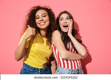 Two Cheerful Young Girls Wearing Summer Clothing Standing Isolated Over Pink Background, Grimacing, Giving Thumbs Up