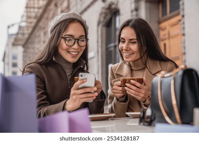 Two cheerful young girls women friends holding smartphone watching photo video using mobile apps texting chatting while sitting at the cafe outdoors urban street - Powered by Shutterstock