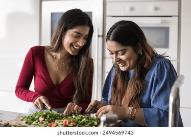 Two cheerful young Indian women chopping fresh vegetables for salad, enjoying domestic cooking activities, family leisure, laughing, having fun at kitchen table, preparing healthy meal for dinner - Powered by Shutterstock