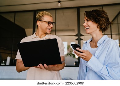 Two cheerful young multinational colleagues talking while standing in office with laptop and mobile phone - Shutterstock ID 2236032909