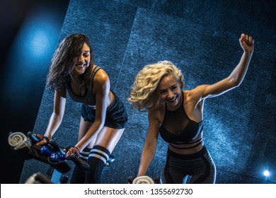 Two cheerful young sporty women doing cardio workout on cycling bikes indoors